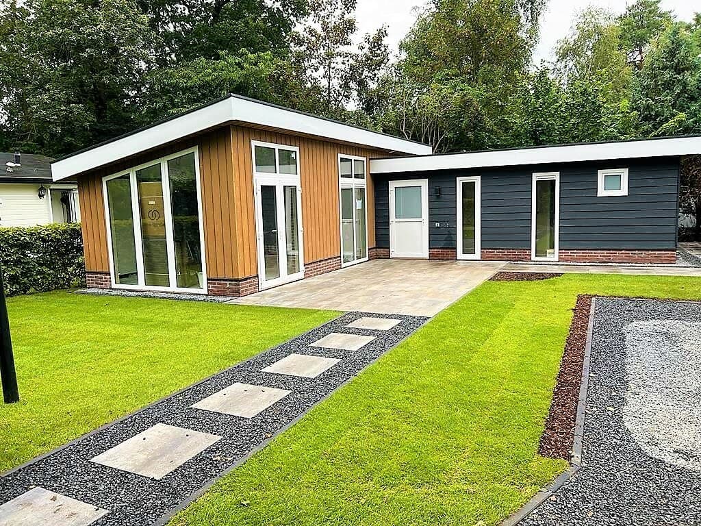 Moderne vakantiewoning met houten en betonnen gevel, grote ramen en een betegeld terras omgeven door groen.