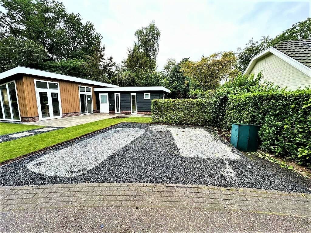 Twee vakantiewoningen met een grasveld en een grindpad, omgeven door hagen en bomen in een rustige omgeving.