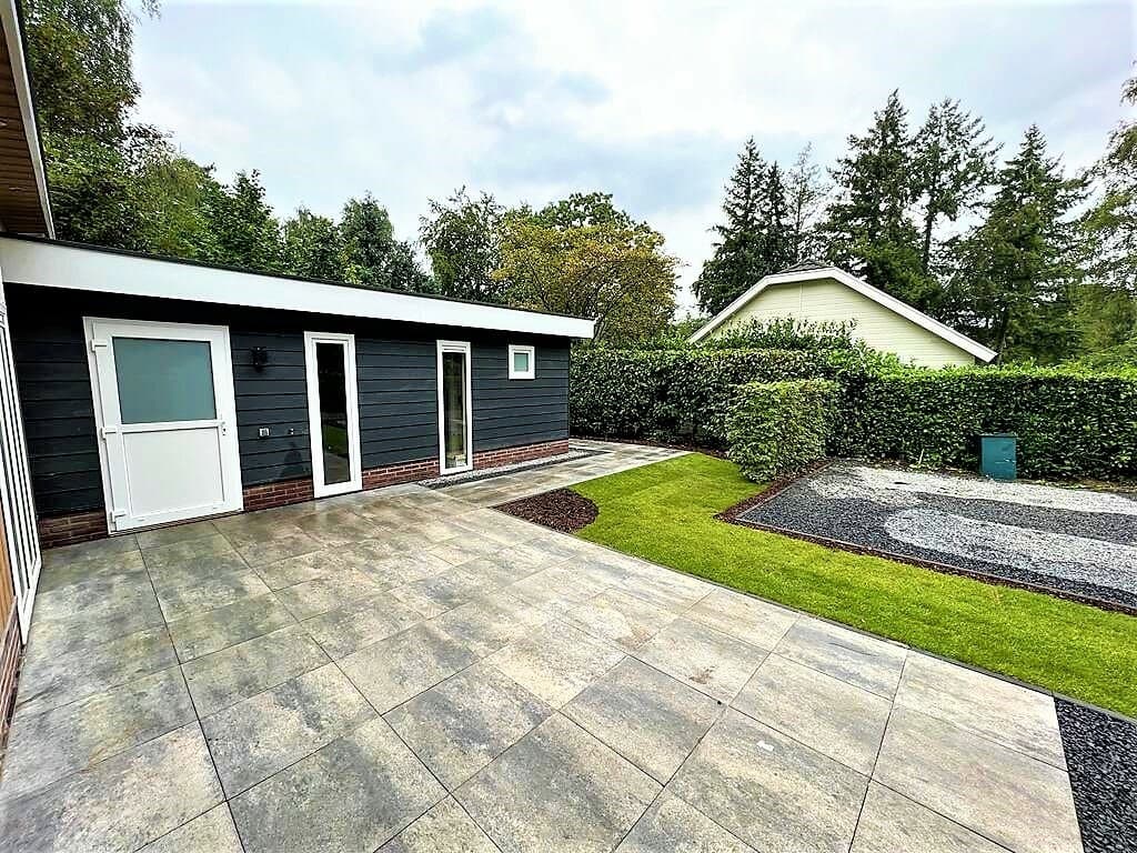 Tegeltuin met groen gras, strakke gevel van een woning en omsloten door hagen en bomen.