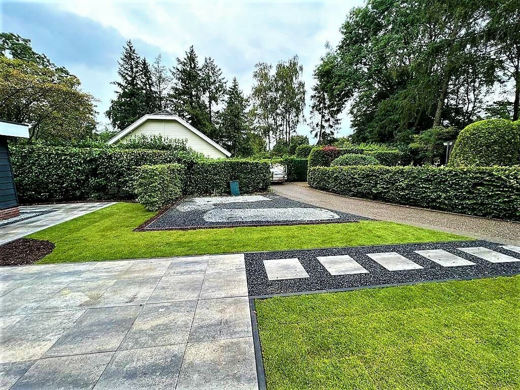 Tuin met groene grasvelden, bestrate paden en hagen, met een parkeerplaats zichtbaar in de achtertuin