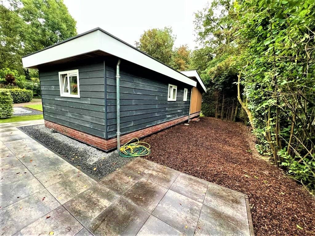 Zijdelingse aanzicht van een woning met houten gevel, omgeven door bomen en een grindpad met verharding.