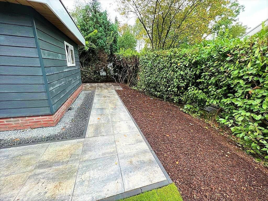 Tuinpad naast een woning omringd door hoge groene hagen en een barkmulchgebied met grind.