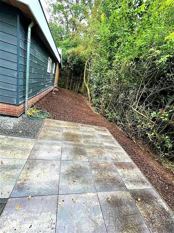 Pad van tegels naast een woning met een groene, bushachtige omgeving en een grindpad aan de zijkant.