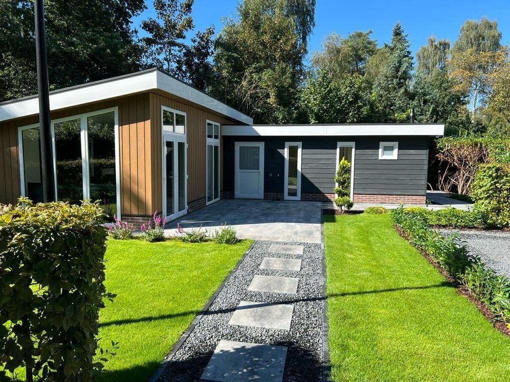 Moderne vakantiewoning met houten panelen en grote ramen, omgeven door groen en een betegeld pad naar de ingang.