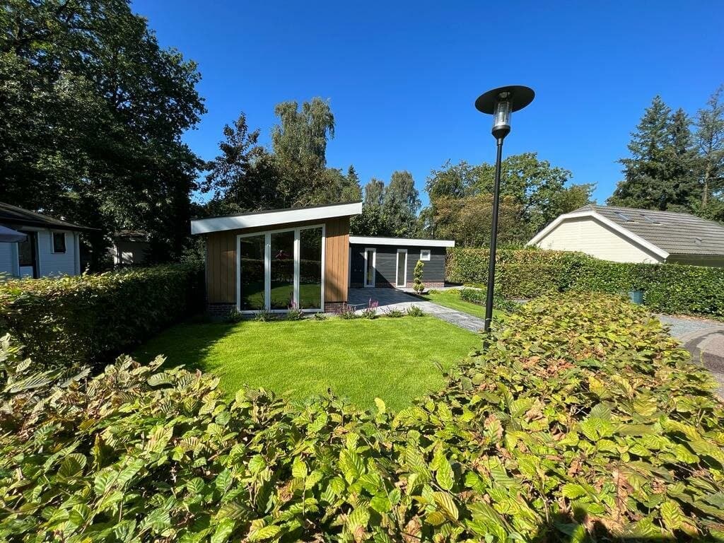 Moderne vakantiewoning met grote ramen, omgeven door groen gras en struiken, met een blauwe lucht op de achtergrond.