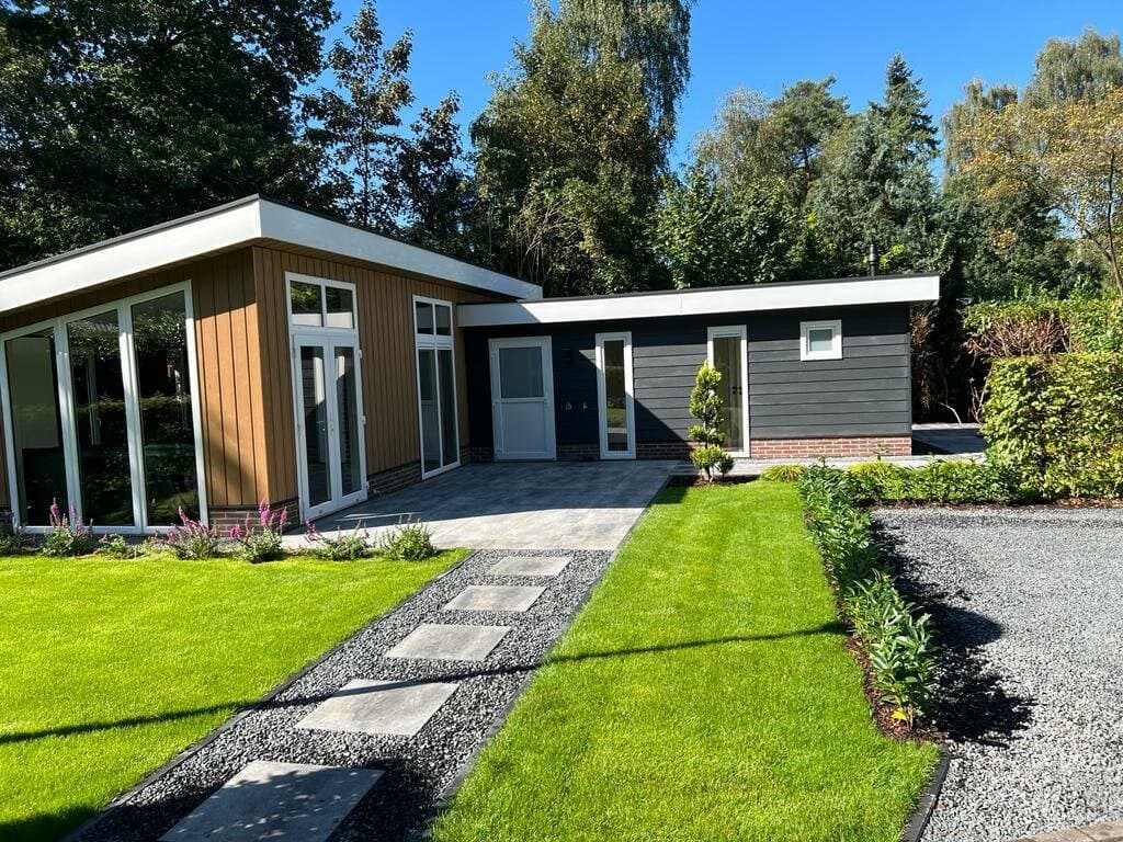 Moderne vakantiewoning met houten en donkere gevel, grasveld en betegelde oprit omgeven door groene beplanting.