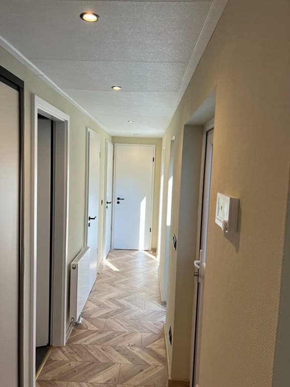 Gang met meerdere deuren, lichte muren, houten vloer en inbouwverlichting aan het plafond