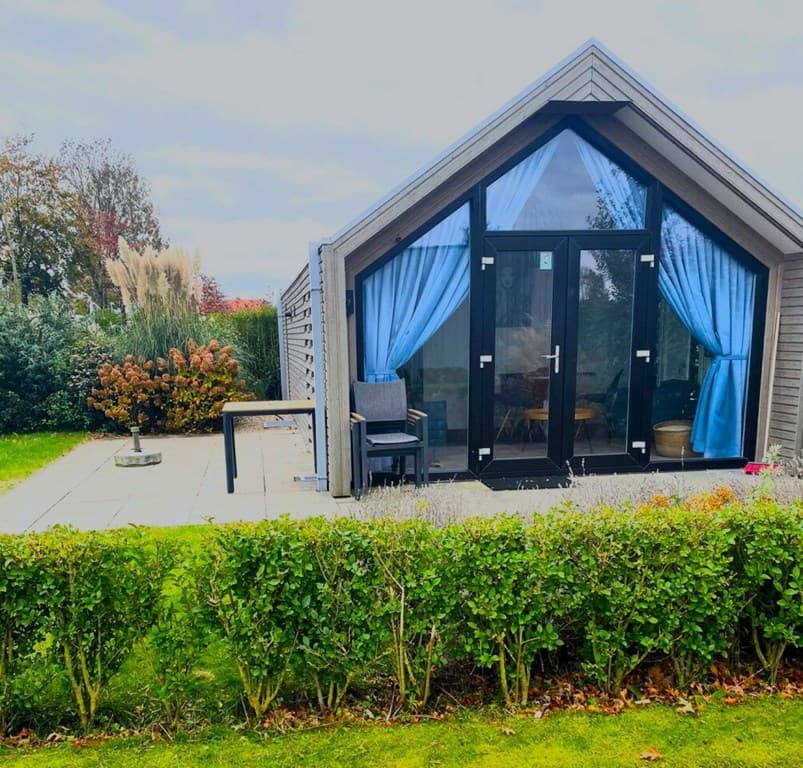 Moderne vakantiewoning met grote glaspartijen, tuinmeubilair en omringende groene hagen in een rustige omgeving