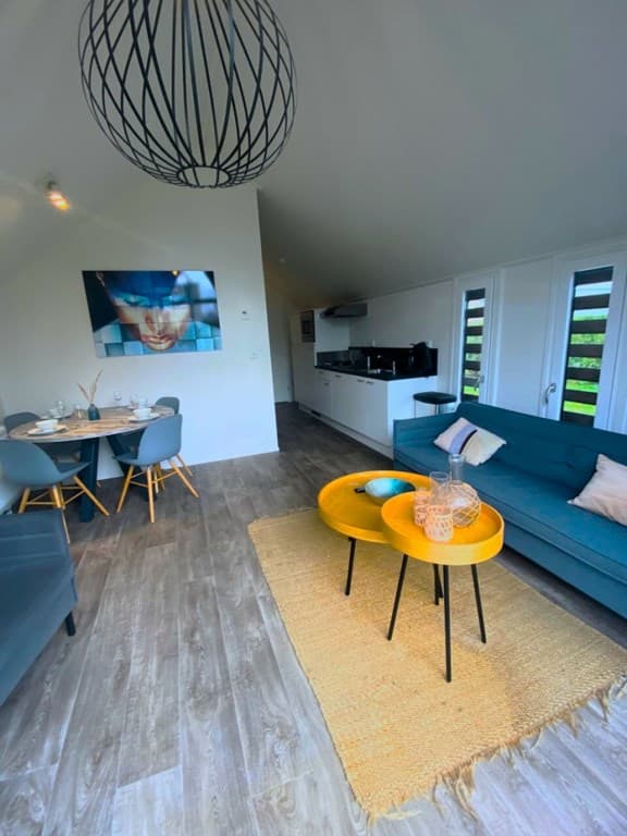 Ruimte met een groene bank, twee ronde tafels, een moderne lamp en een open keuken in een lichte omgeving.