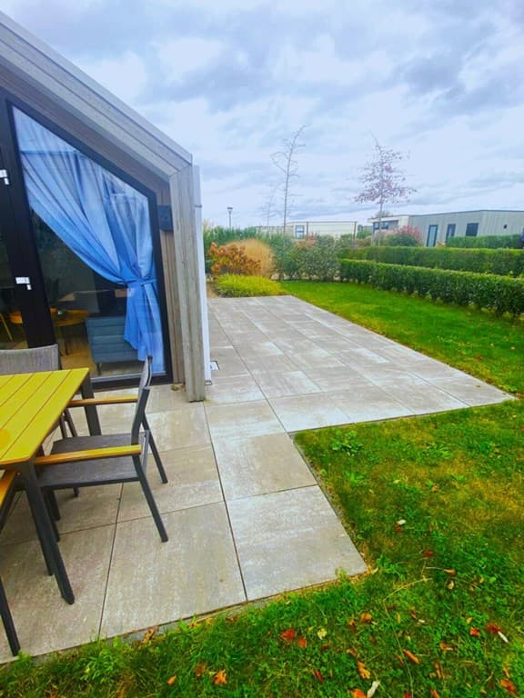 Terras met houten tafel en stoelen, gazon en groene heggen rondom de woning, gedeeltelijk overdekt door een blauwe gordijn.