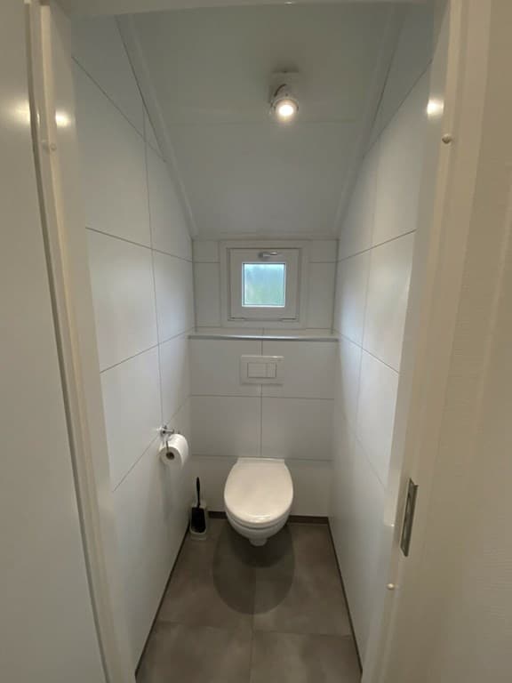Klein toilet met witte wandtegels, raam en uitzicht op een lichte tegelvloer.