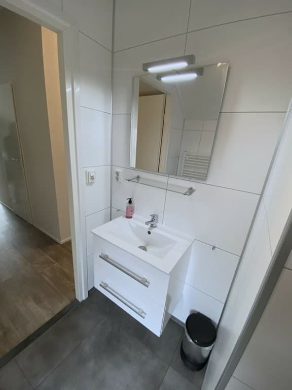 Badkamer met witte wandtegels, een wastafel met spiegel en verlichting, en een deur naar een andere ruimte.