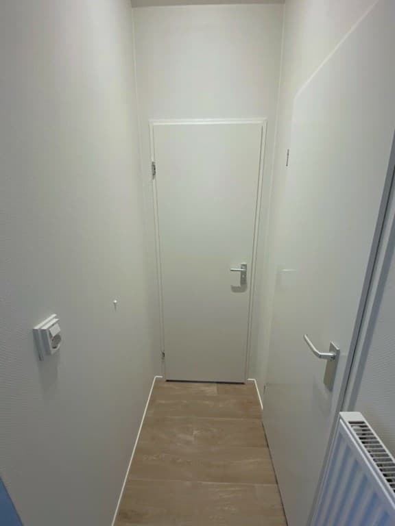 Smal gang met lichte wanden, een deur aan het einde en een radiator aan de rechterzijde.