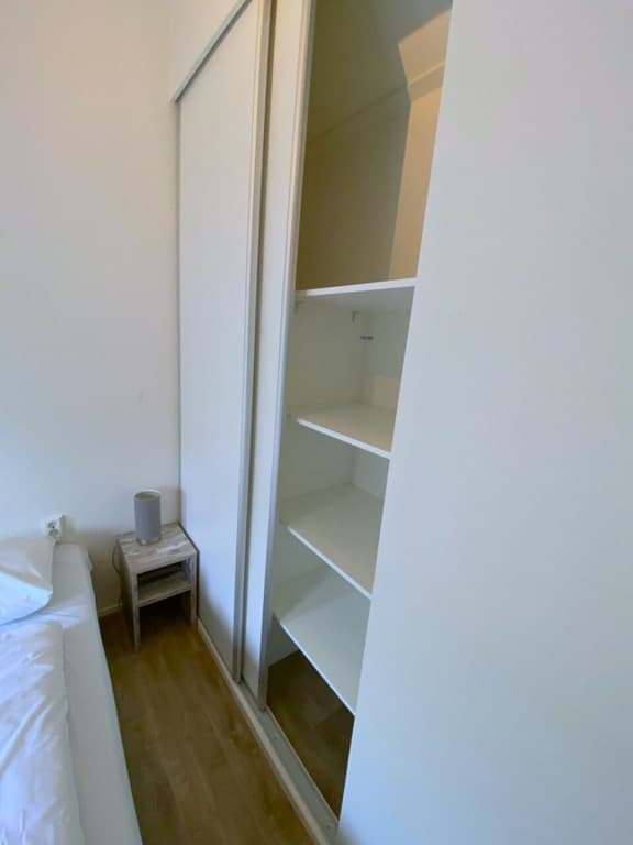 In een kamer met een witgekleurd interieur, een kast met planken en een bed met witte beddengoed zichtbaar.
