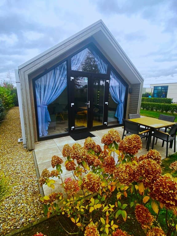 Vakantiehuis met moderne gevel, grote ramen en een overdekte patio, omringd door bloeiende planten en gras.