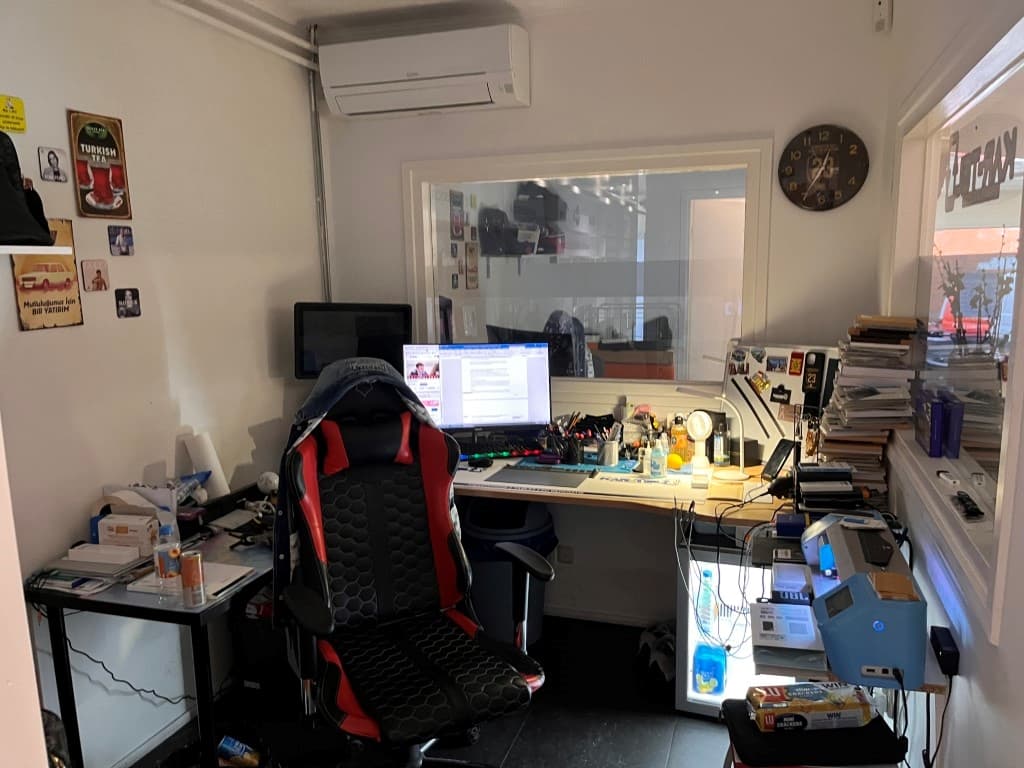Kantoorruimte met een bureau, een computerscherm, een bureaustoel en verschillende stapels papieren en tijdschriften.