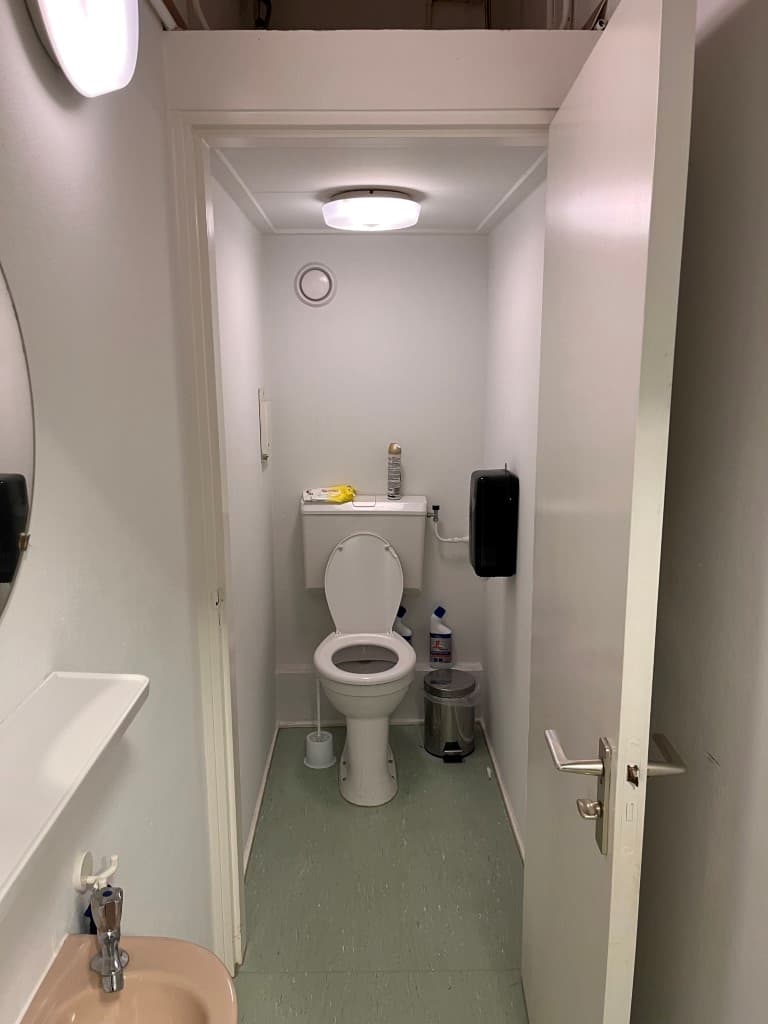 Ingang van een sanitaire ruimte met toilet en wastafel, lichtgrijze muren en groene vloer.