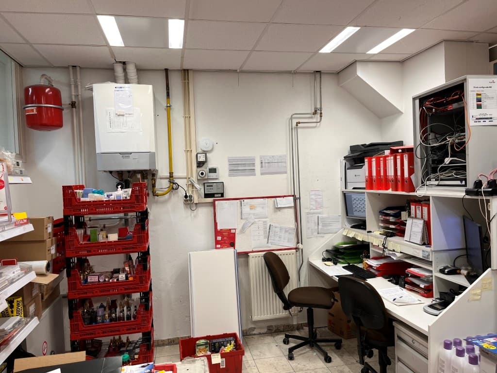 Kantoorruimte met bureau, kasten en diverse opslagdozen tegen de wand en een verwarmingsinstallatie aan de muur.
