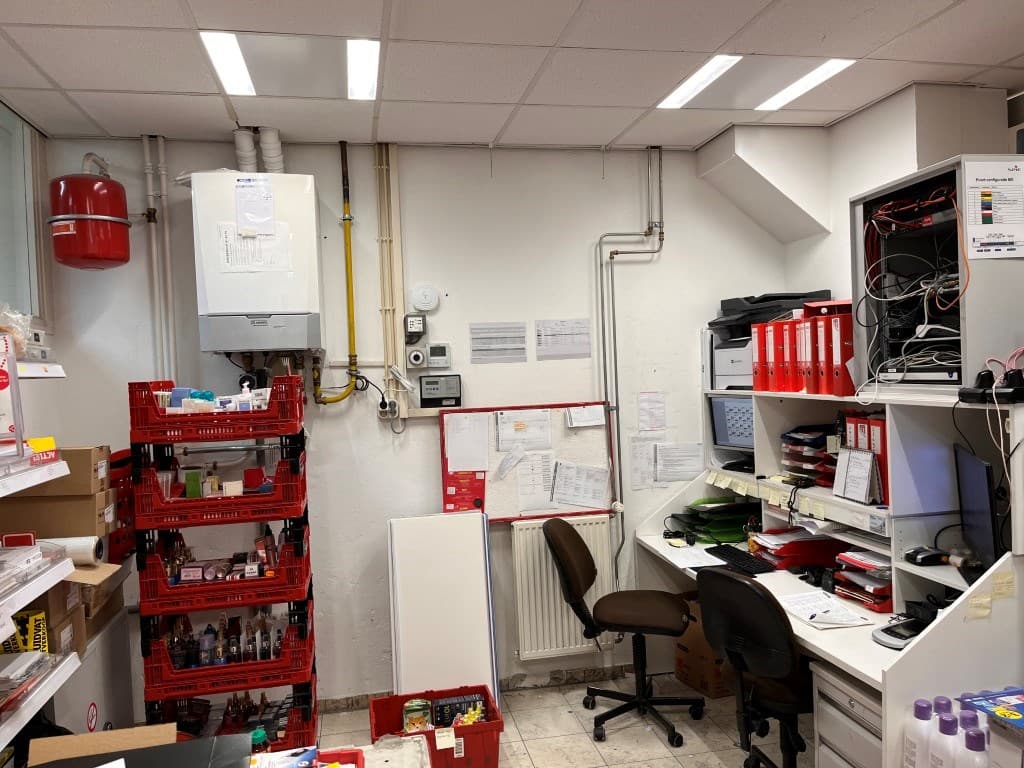 Kantoorruimte met bureau, kasten en diverse opslagdozen tegen de wand en een verwarmingsinstallatie aan de muur.