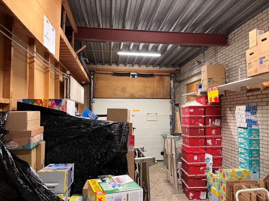 Opslagruimte met gestapelde dozen, plastic bakken en een rolluik, verlicht door TL-verlichting aan het plafond.