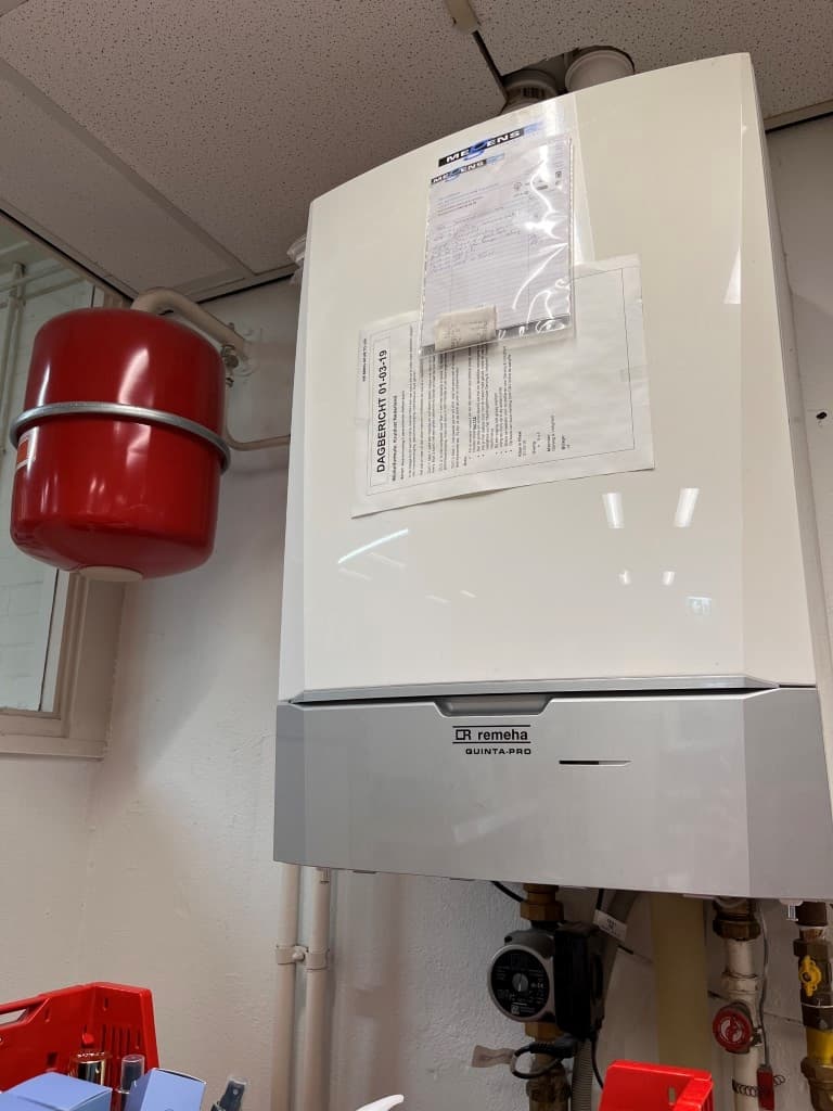 Huiselijke ruimte met een witte ketel, een rode expansietank en verschillende leidingen zichtbaar aan de muur.