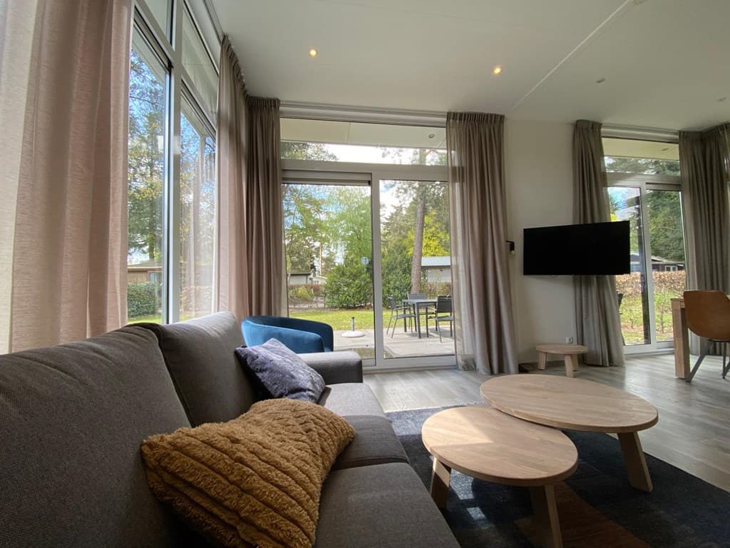 Moderne woonkamer met grote ramen, grijze bank, houten tafels en uitzicht op groen buiten met terras set.