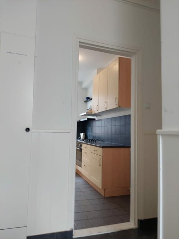 Keuken met houten kasten, een fornuis en donkere wandtegels, zichtbaar vanuit een hal met lichte muren