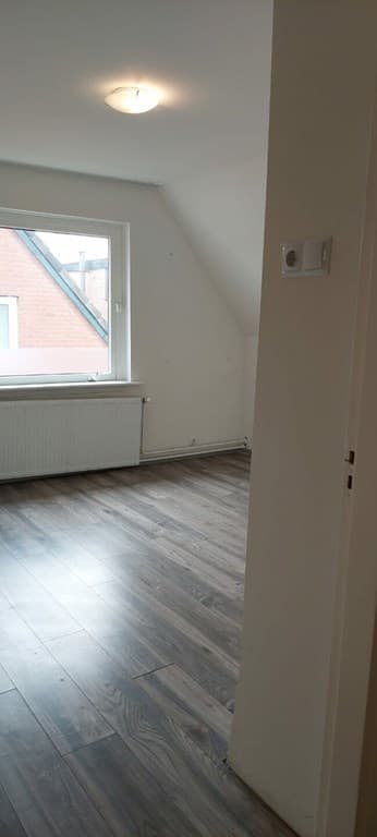 Ruimtelijke woonkamer met laminaatvloer, groot raam en zicht op de wand en deur naar de gang.