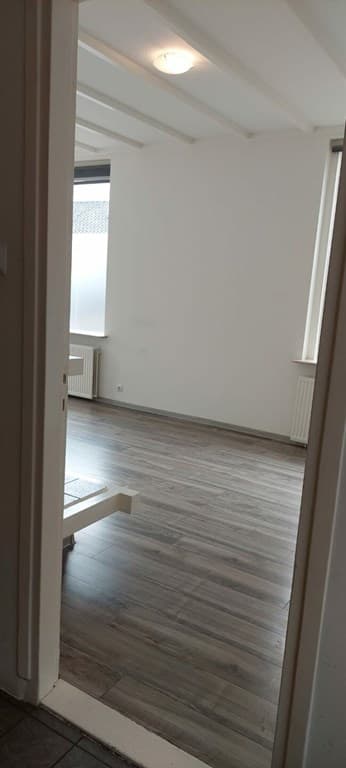 Kamer met kunststof vloeren, witte muren en een raam met lichtinval, zichtbaar vanaf een gangdeur.