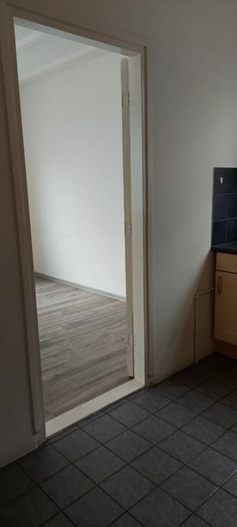De foto toont een doorgang naar een lichte kamer met een houten vloer en een keukenhoek met enkele keukenkastjes.