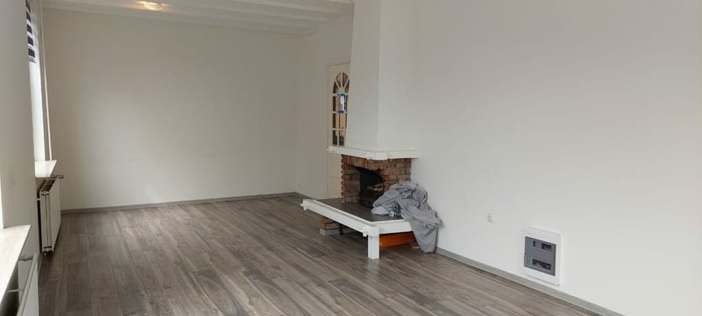 Woonkamer met houten vloer, open haard van steen en een eenvoudige muurkachel.