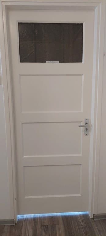 Witte binnendeur met een venster bovenin en een eenvoudige deurknop, tegen een lichte wand geplaatst.