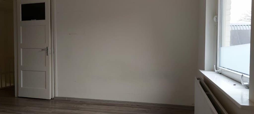 Lege kamer met witte muren, een raam, radiator en deur in een nette afwerking met houten vloer.