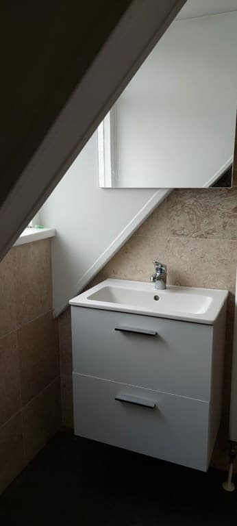 Badkamer met wastafel en spiegel onder een schuin plafond, betegelde wand en vloer met donkere tegels