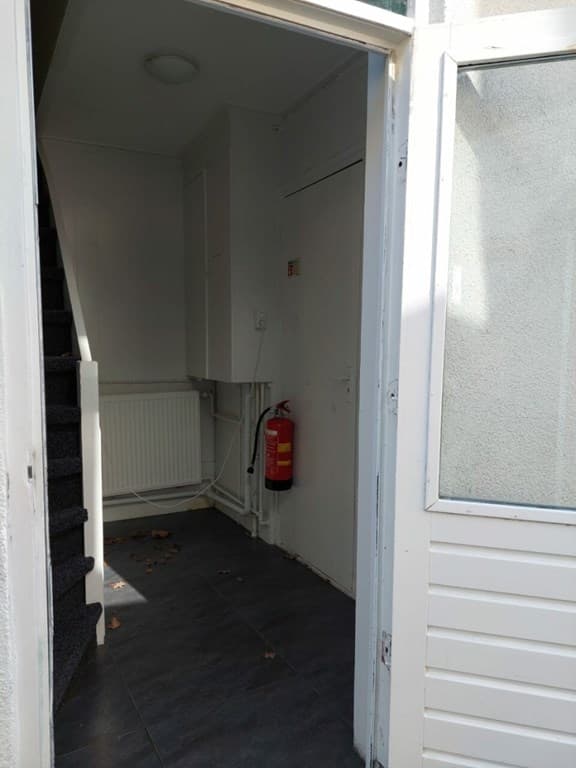 Entree met trap, radiator en brandblusser aan de muur, vloer van tegels en witte wandafwerking.