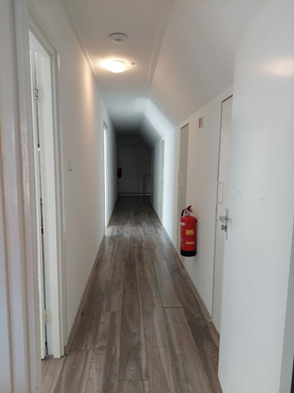 Langgerekte gang met houten vloer en witte deuren aan beide zijden, lichtinval van boven en een brandblusser aan de muur