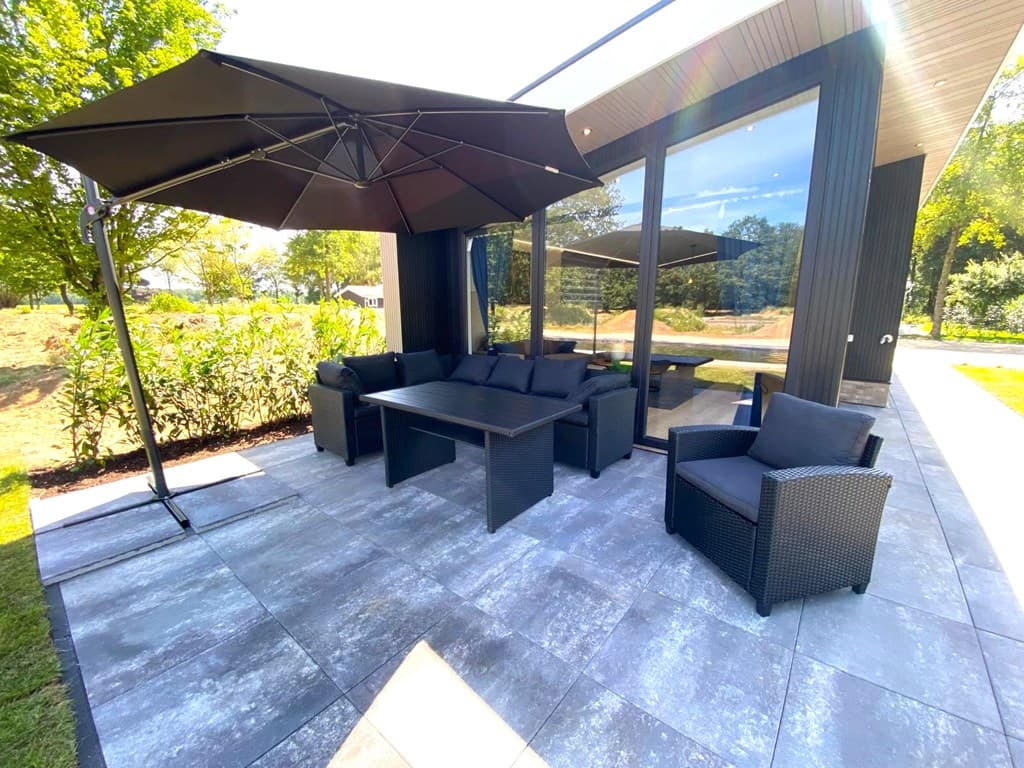 Terras met een loungebank, stoelen en parasol, omgeven door groene begroeiing en grote ramen van de woning