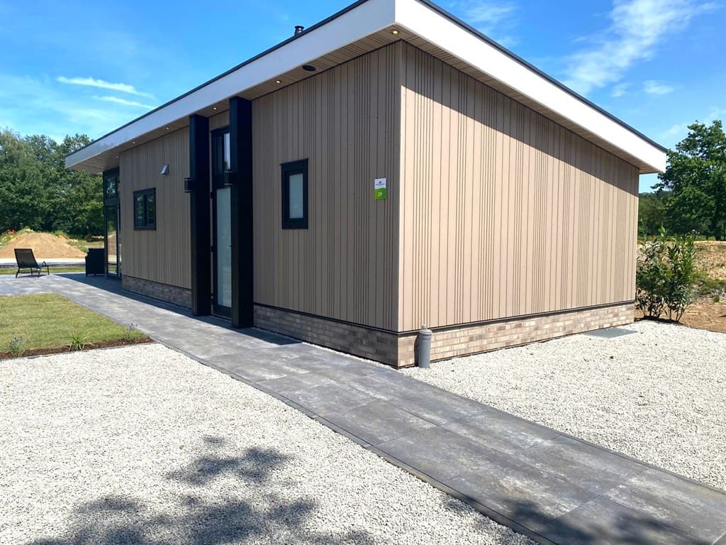 Moderne vakantiewoning met houten gevel, grote ramen en een betegeld pad naar de ingang, omgeven door een gazon.