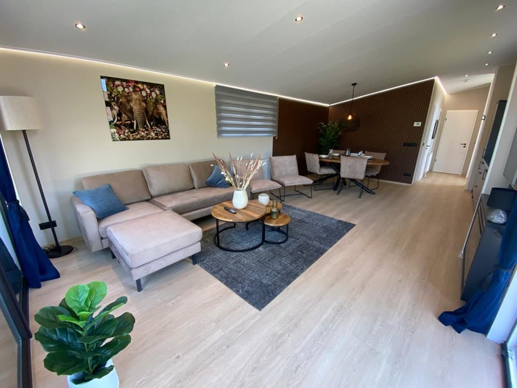 Ruime woonkamer met een lichte bank, salontafel, eetkamertafel en een decoratieve muur met kunstwerk en planten