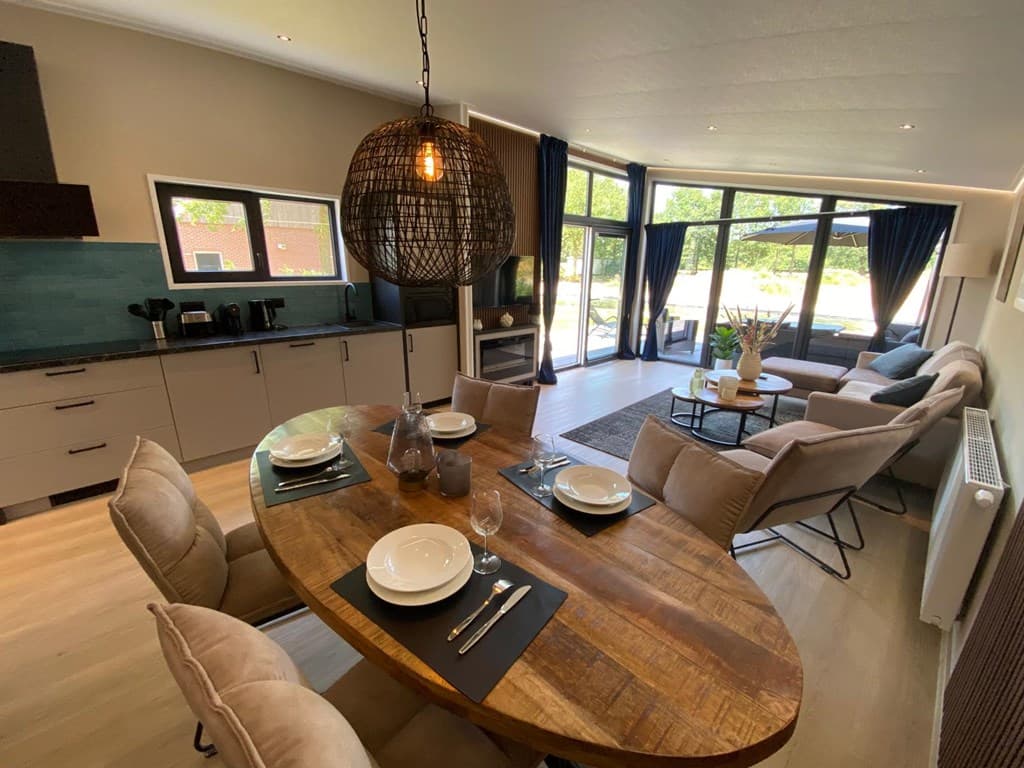 Modern ingerichte woonkamer met open keuken, grote ramen, eettafel met stoelen en zithoek met bank en fauteuils.