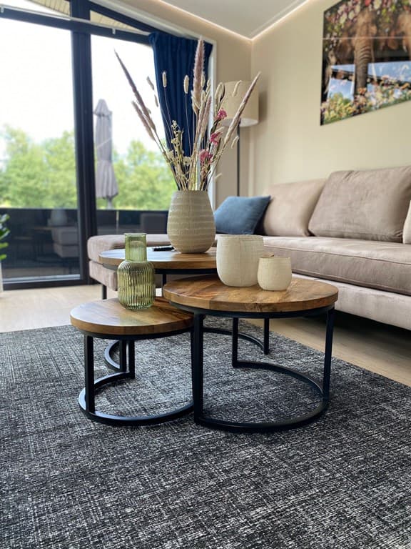 Woonkamer met een bank, salontafels van hout en metaal, decoratieve vazen en een groot raam met uitzicht op groen.