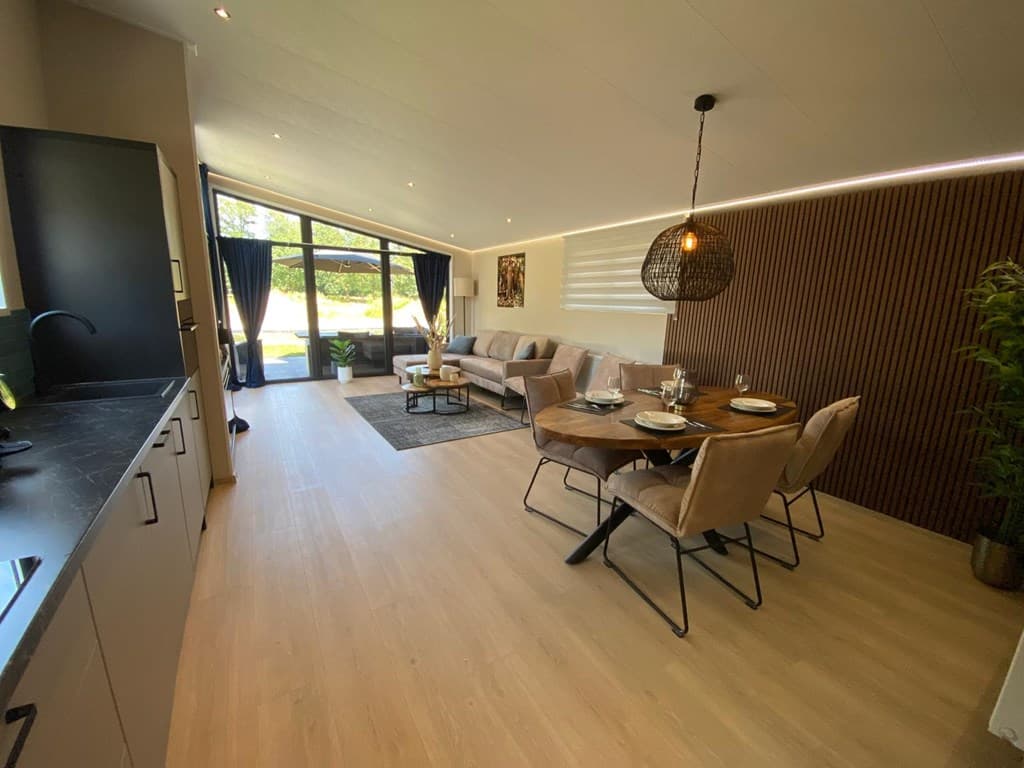 Moderne woonkamer met open keuken, eettafel en ruime zithoek met grote ramen en uitzicht op de tuin.