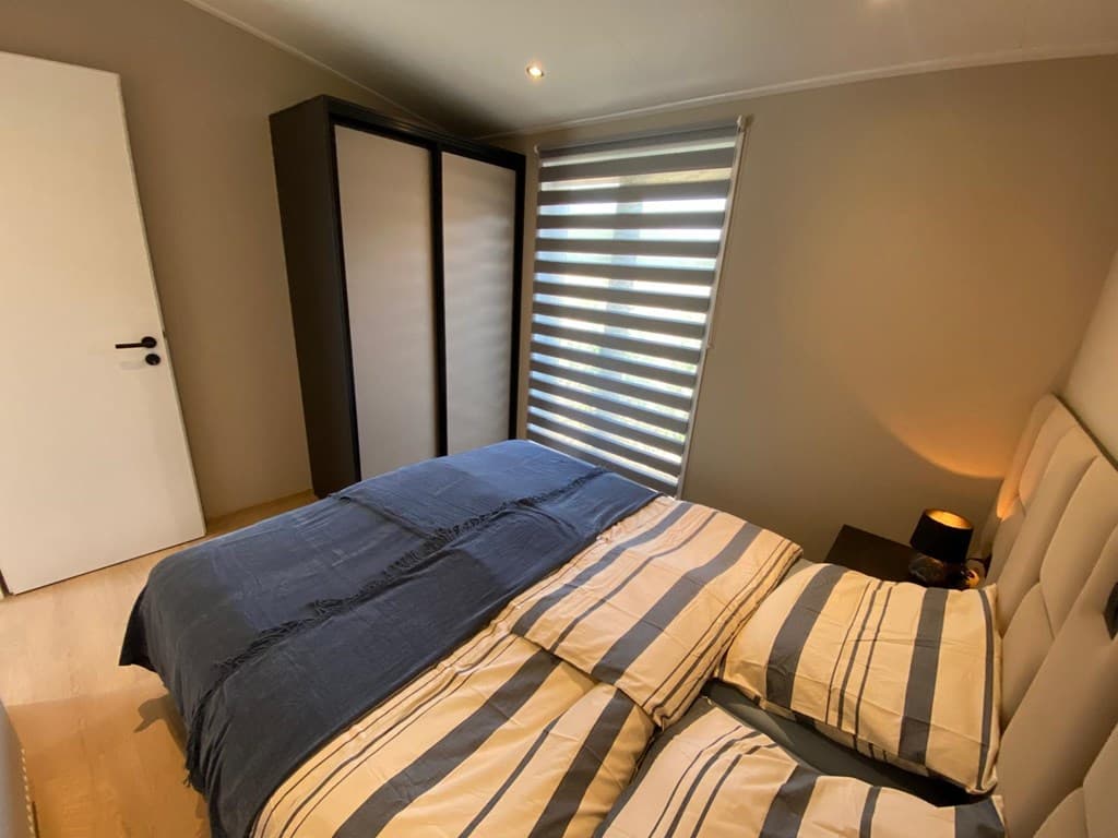 Slaapkamer met een tweepersoonsbed, nachtkastje en een strakke kast naast een raam met jaloezieën.