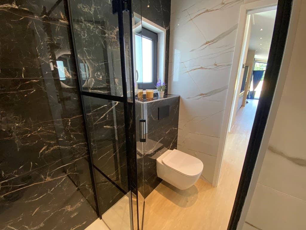 Moderne badkamer met een inloopdouche, wand met marmerlook en een toilet, met zicht op een lichte kamer erachter.