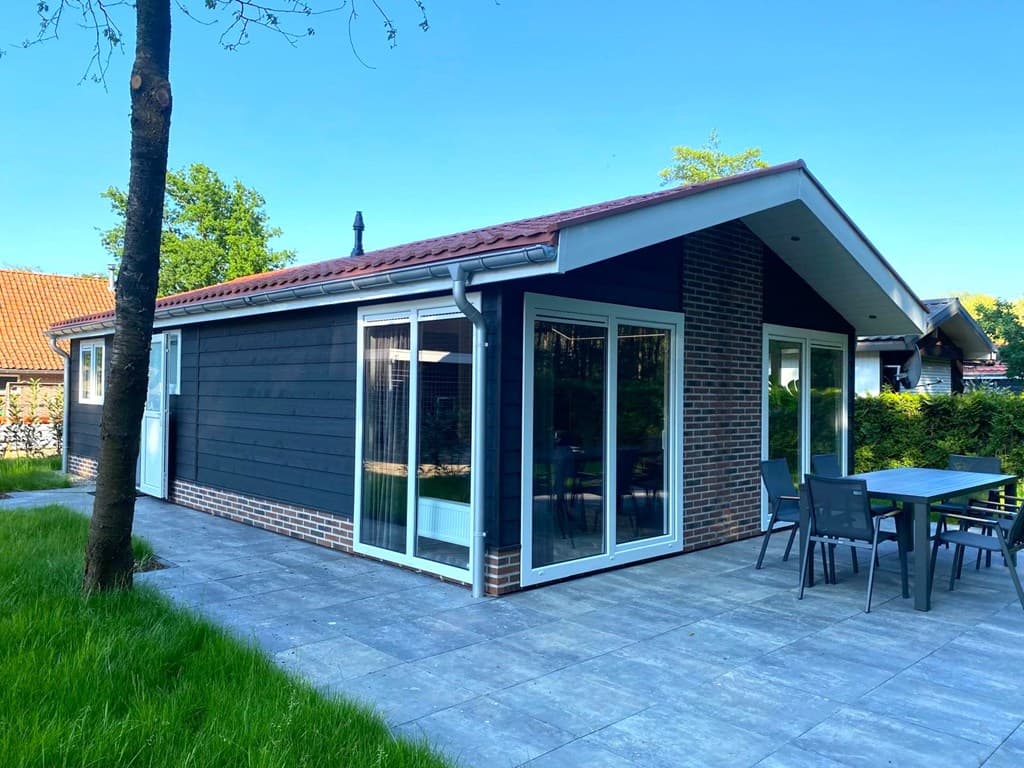 Moderne vakantiewoning met grote ramen, terras en tuinmeubilair op een groene locatie.