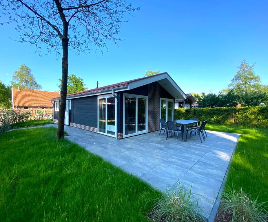 Moderne vakantiewoning met grote ramen, een betegeld terras en grasvelden rondom, gelegen in een groene omgeving.