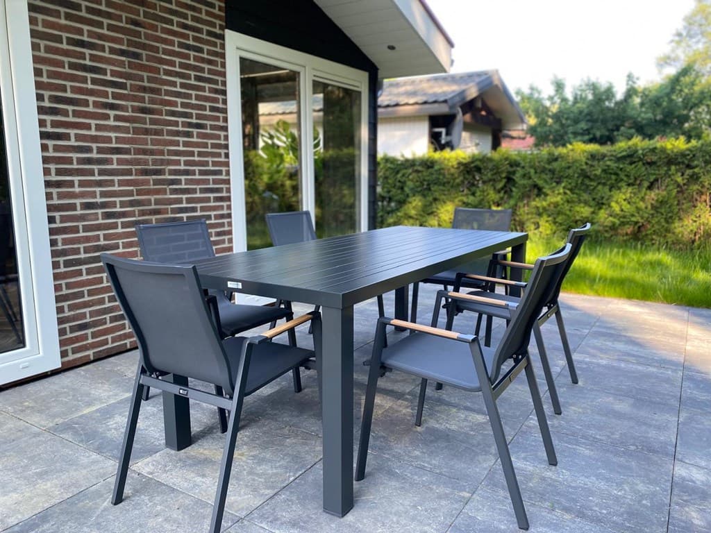 Tuinset met een lange tafel en zes stoelen op een betegeld terras bij een woning met een groene haag op de achtergrond