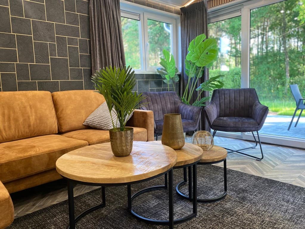 Moderne woonkamer met een bruine bank, houten salontafels en een groot raam met uitzicht op de tuin, planten in de hoek.