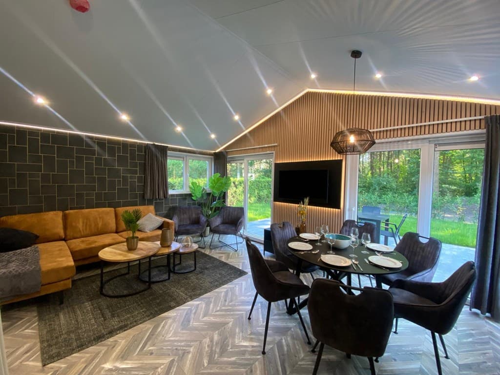 Moderne woonkamer met een zithoek, grote ramen en een eethoek met acht stoelen, omringd door natuur.