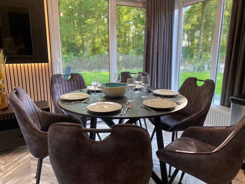 Eettafel met zes stoelen, servies en bestek, met uitzicht op groene natuur door grote ramen.